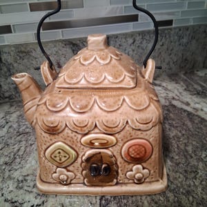 May include: A tan ceramic teapot shaped like a house, featuring a black wire handle and decorative details. The teapot has a scalloped roof, a small door, and various embossed shapes and floral accents. The spout and lid are also visible.