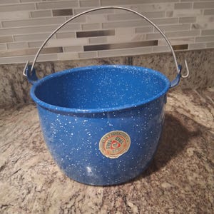 May include: A blue enamelware bucket with white speckles and a silver wire handle. The bucket has a round shape and a label with text. This vintage bucket is suitable for camping or home decor.
