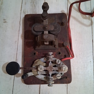 May include: A vintage telegraph key with a wooden base and metal components. The key has a lever with a black knob and a set of metal contacts. The key is connected to a red wire.