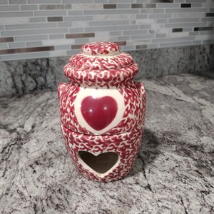 May include: A white ceramic jar with a red heart design. The jar has a lid and a cutout heart on the bottom. The jar is decorated with a red and white speckled pattern.