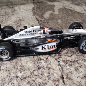 May include: A black and silver Formula 1 race car with the words "Mobil 1", "Mercedes Benz", "Kimi", and "Siemens" on the side. The car is on a gray and white marble surface.