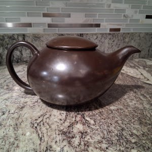 May include: A dark brown ceramic teapot with a rounded body and a curved spout. The teapot has a smooth, glossy finish.