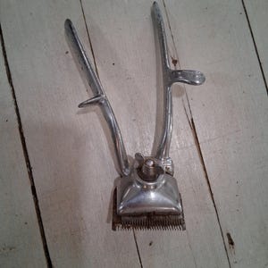 May include: A vintage silver hair clipper with a handle and a blade. The clipper has a small knob on the top.