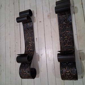 May include: Two black metal wall sconces with a scroll design. Each sconce features a decorative pattern in a bronze color and a holder for a single candle. The sconces are mounted on a white wooden surface.