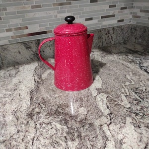 May include: A red speckled enamel coffee pot with a black lid and handle. The pot is sitting on a gray and white countertop.