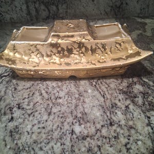 May include: A rectangular ceramic planter with a metallic gold finish featuring a textured, mottled surface. The vessel has a raised central section and two open compartments on either side, with a cream-colored interior visible throughout.