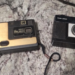 May include: Two vintage cameras are displayed on a textured surface. One is a Kodak Disc 3100 with a gold and black design. The other is a black camera with the text "EAS-LOAD" and "185-PF". Both cameras have a strap.