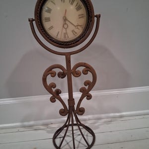 May include: A decorative, freestanding clock with a round face and Roman numerals. The clock is set in a rusty brown metal frame with a heart-shaped base and ornate scrollwork. The clock's hands point to approximately 2:30.