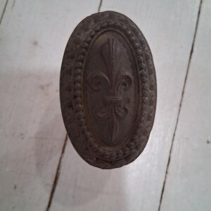 May include: Oval-shaped, antique bronze-colored decorative hardware with a fleur-de-lis design. The hardware features a beaded border and is likely a cabinet knob or drawer pull.