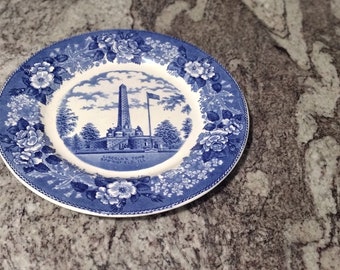 May include: A blue and white ceramic plate with a floral design. The center of the plate features a black and white image of a building with a tall tower and a flag.