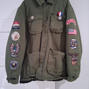May include: Olive green jacket with multiple patches and a small medal. Patches include an American flag, an eagle, and the words "LAND OF THE FREE." The jacket has four front pockets and a button-up closure. The jacket is hanging on a green hanger.