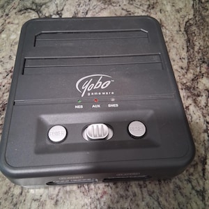May include: A dark gray Yobo Gameware console with power and reset buttons. The console has slots for NES, AUX, and SNES cartridges. The Yobo Gameware logo is visible on the front of the console.