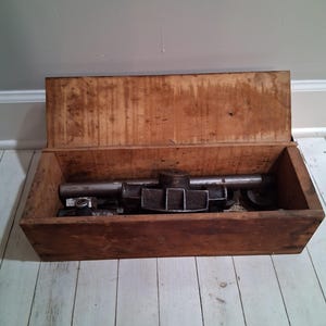 May include: A wooden box with a hinged lid, open to reveal a set of vintage metal pipe tools. The tools include a pipe wrench, a pipe cutter, and a pipe threader.
