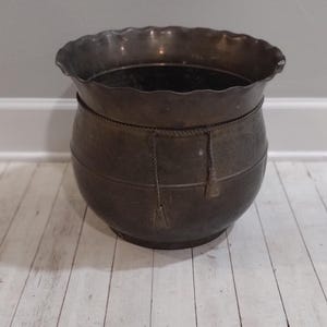 May include: A dark brown metal planter with a scalloped rim and rope accents. The planter has a distressed finish and is sitting on a white wood floor.
