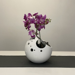 May include: A white ceramic vase with a moon-like design and holes throughout. The vase is filled with a bouquet of purple orchids.