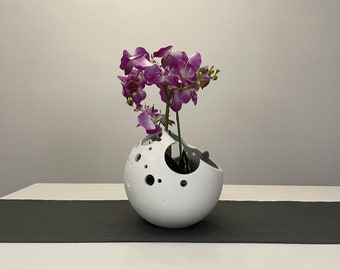 May include: A white ceramic vase with a moon-like design and holes throughout. The vase is filled with a bouquet of purple orchids.