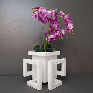 May include: White ceramic vase with a square base and four open square frames extending from the sides. The vase is filled with a pink orchid plant.