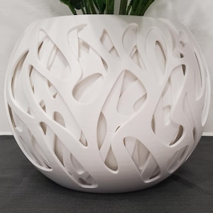 May include: A white, spherical decorative vase with an open, interwoven design. The vase is filled with green plants and sits on a dark gray surface. The vase's intricate pattern creates a modern, artistic look.