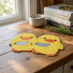 May include: Two yellow chick-shaped coasters with orange feet and beaks, each holding a gray knife. The coasters are embroidered with black eyes and are placed on a wooden surface. A small ceramic bowl and books are in the background.