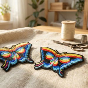 May include: Two embroidered butterfly patches with vibrant rainbow colors on a neutral linen surface. The patches feature intricate details and are surrounded by sewing tools, including scissors, thread, and a thimble. The patches are approximately 4 inches wide.
