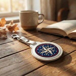 May include: A white leather keychain with a compass design. The compass has a navy blue background with a gold star and white dots, framed by a red and white border. It is on a wooden surface.