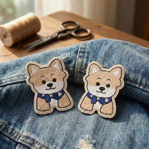 May include: Two embroidered patches featuring a cartoon Shiba Inu dog. The patches have tan fur, white accents, and a blue collar. They are placed on a denim jacket. Sewing supplies, including scissors and thread, are in the background.