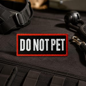 Pode incluir: Um emblema retangular bordado com fundo preto e uma borda vermelha vibrante. O texto DO NOT PET está costurado em letras maiúsculas brancas no centro do emblema. O design apresenta uma estética limpa e de alto contraste.