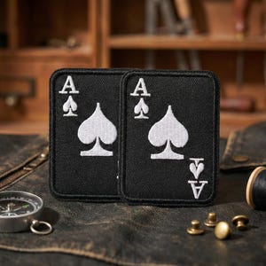 May include: Two black Ace of Spades patches with white spade symbols and the letter "A". The patches are rectangular and have a stitched border. The patches are displayed on a textured, dark surface.