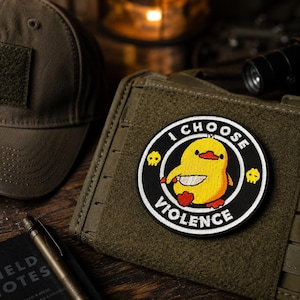 Puede incluir: Un parche bordado circular que presenta un patito amarillo sosteniendo un cuchillo, rodeado por un borde negro con el texto blanco I CHOOSE VIOLENCE y dos pequeños iconos de calaveras amarillas. El parche está sujeto a un panel táctico verde oliva.