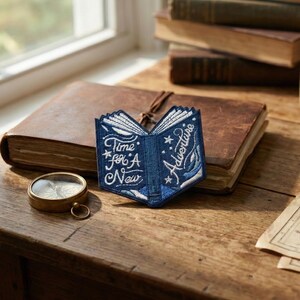 以下が含まれることがあります： 「Time for A New Adventure」の文字が入った、開いた本型の青い刺繍パッチ。革張りのジャーナルとコンパスの隣の木製の表面にパッチが置かれています。