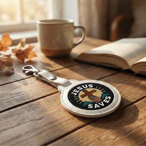 May include: White leather keychain with a silver clasp. The round emblem features the text "JESUS SAVES" around a cross, hand, and clouds design. The keychain is on a wooden surface.