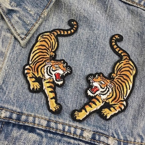 May include: Two embroidered tiger patches with orange and black stripes, white accents, and open red mouths. The patches are on a blue denim background.