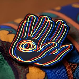 May include: A black patch shaped like an open hand with a central eye symbol. The design features layered, neon-colored outlines in red, blue, and yellow, creating a vibrant, multi-dimensional effect against the solid black base of the patch.