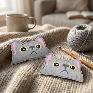 May include: Two gray cat-shaped patches with pink ears and yellow eyes are displayed on a beige knitted blanket. A ball of gray yarn and a wooden crochet hook are also visible. A beige mug and a couch are in the background.