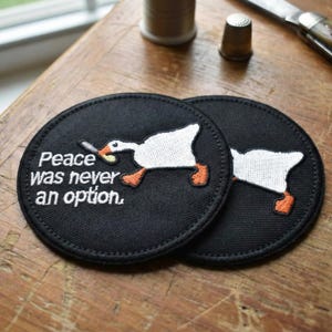 Puede incluir: Dos posavasos redondos negros con un diseño de ganso blanco y el texto "Peace Was Never an Option". El ganso se representa con un cigarrillo. Los posavasos están sobre una superficie de madera.