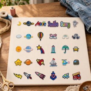 May include: A collection of twenty-five embroidered iron-on patches arranged in a grid on a white fabric board. The designs include planets, stars, vehicles, weather icons, and the words Music, Smile, and Sunny in colourful thread.