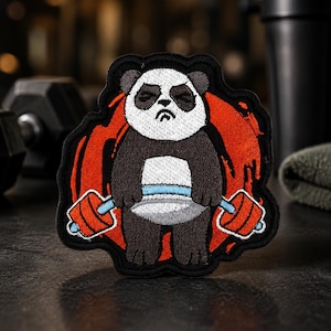 Puede incluir: Un parche bordado que muestra un panda gruñón de pelaje blanco y negro, sosteniendo una barra de pesas con discos rojos y una barra azul claro. El panda está centrado sobre un fondo circular rojo texturizado con un borde negro grueso.