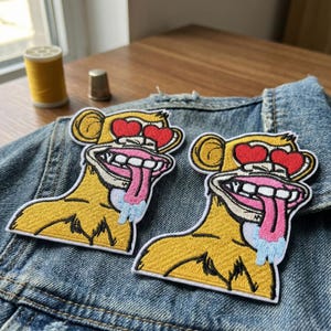 May include: Two embroidered patches of a cartoon monkey with heart-shaped eyes and a long tongue. The monkey is yellow with a white border, and the tongue is pink with blue drool. The patches are on a denim jacket.