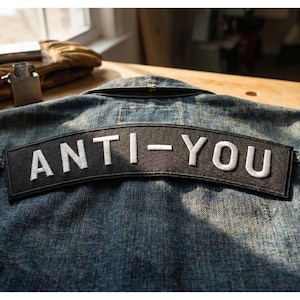 Puede incluir: Chaqueta vaquera con un parche negro bordado con las palabras "ANTI-YOU" en letras blancas. El parche tiene forma de arco y está cosido en la parte trasera de la chaqueta.