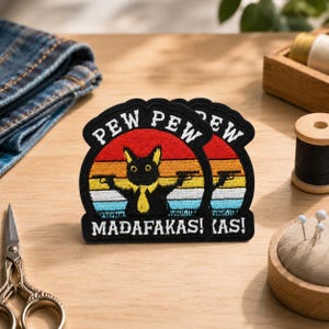 Peut inclure: Un écusson brodé représentant un chat noir portant une cravate jaune et tenant deux pistolets. Le fond est un motif de coucher de soleil avec des bandes rouges, orange, jaunes, blanches et bleues. Le texte indique PEW PEW MADAFAKAS! en blanc.