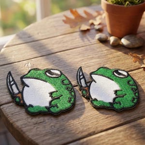 May include: Two embroidered patches featuring a green frog holding a knife. The frog has white and black details, with a black outline. The patches are on a wooden surface.
