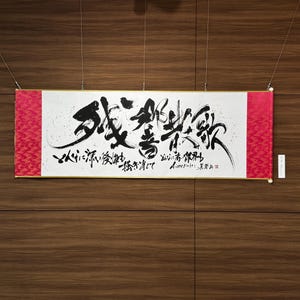 May include: A rectangular Japanese calligraphy scroll with black ink characters on a white background. The scroll has red patterned end pieces and is suspended by wires against a wood-paneled wall. The calligraphy includes smaller text below the main characters.