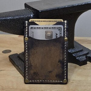 May include: A handcrafted, brown leather card wallet with visible stitching and brass rivets. The wallet holds several credit cards. The background includes a black anvil and a wooden surface, suggesting a focus on craftsmanship and durability.