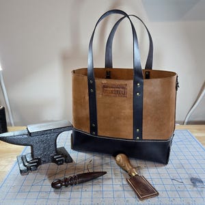 May include: A handcrafted leather tote bag with a brown body and black base and accents. The bag features sturdy handles and is displayed with leatherworking tools, including an anvil and a wooden handled knife. The bag has the text "Hand Made Goods" on it.