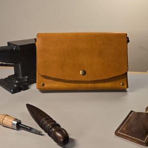 May include: A handcrafted, tan leather bag with a flap closure and brass hardware. The bag is displayed with leatherworking tools, including a stitching awl, a burnisher, and a leather stamping tool. An anvil is in the background.