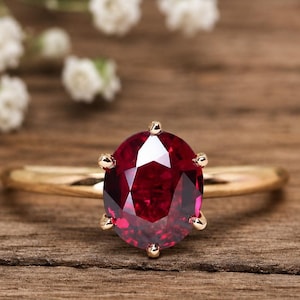 Peut inclure: Une bague en or ornée d'un grand rubis ovale. Le rubis, d'un rouge profond, est serti de six griffes. La bague repose sur une surface en bois, avec des fleurs blanches en arrière-plan.