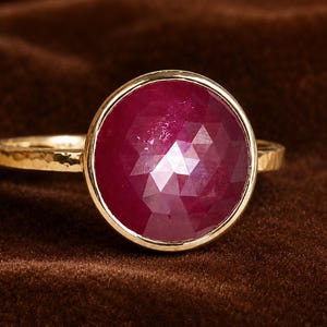 May include: A gold ring with a large, faceted ruby gemstone. The ruby is a deep red colour and is set in a gold bezel. The ring has a hammered gold band.