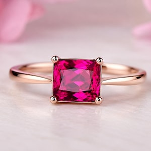 Pode incluir: Um anel de ouro rosa com uma vibrante pedra preciosa de rubi de corte quadrado. O rubi é de cor rosa profundo, cravejado com quatro garras. A banda é de ouro rosa liso e polido. O fundo é rosa e branco suave e desfocado.