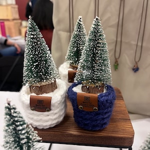 Könnte beinhalten: Drei kleine künstliche Weihnachtsbäume in weißen und blauen gehäkelten Töpfen mit braunen Lederetiketten, auf denen "Made by Sidney Lane Creations" steht.