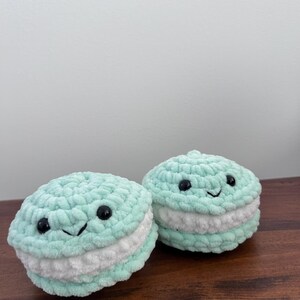 May include: Two crocheted macarons with smiling faces. The macarons are light green with white filling.